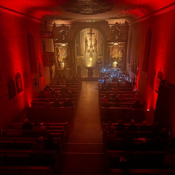 Blick von der Empore auf die Prosselsheimer Kirche, die mit roten Strahlern erleuchtet ist. Der Altarraum ist golden bestrahlt.