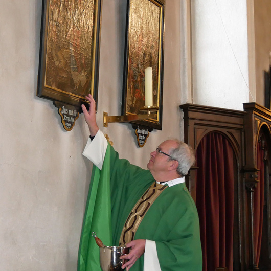 Der Pfarrer hat ein Weihwassergefäß in der Hand und schaut zu zwei Bildern hoch, die an der Wand hängen. Mit der rechten Hand berührt er eines der Bilder.