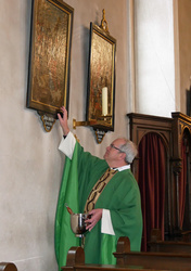 Der Pfarrer hat ein Weihwassergefäß in der Hand und schaut zu zwei Bildern hoch, die an der Wand hängen. Mit der rechten Hand berührt er eines der Bilder.