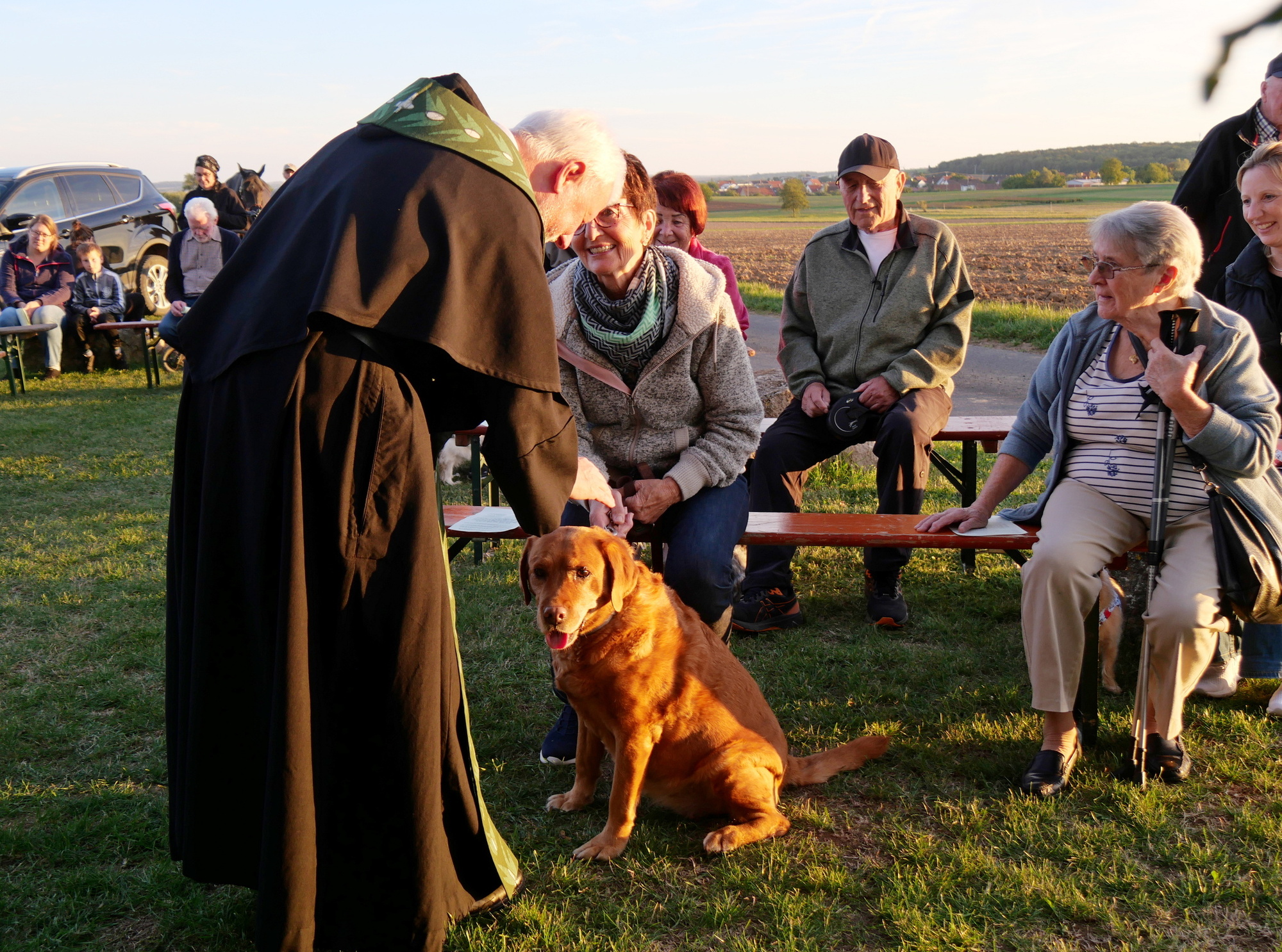 Das Bild zeigt einen Pater, der sich zu einem Hund hinunterbeugt und ihn streichelt. Um ihn herum sitzen ein paar Menschen und schauen ihm zu. Die Stimmung ist friedlich und es ist sonnig.