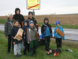 Das Bild zeigt eine Gruppe von neun Kindern, die an einer Straße stehen. Sie halten selbstgebaute Holzgegenstände und tragen wetterfeste Kleidung. Im Hintergrund ist ein Wegweiser mit Ortsnamen und Entfernungen zu sehen. Die Umgebung ist ländlich mit Feldern und einer Straße.