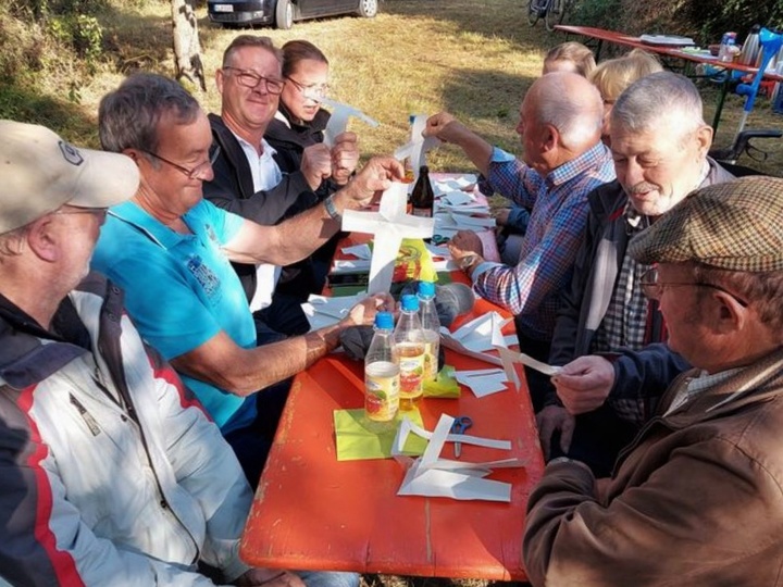 Menschen sitzen um einen Biertisch und schneiden aus weißem Papier Kreuze aus.